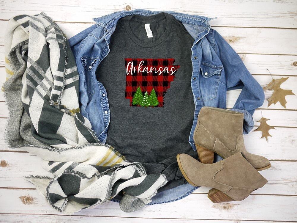 The state of Arkansas in a red plaid silhouette with Arkansas written across in white and Christmas trees at the bottom of the state.